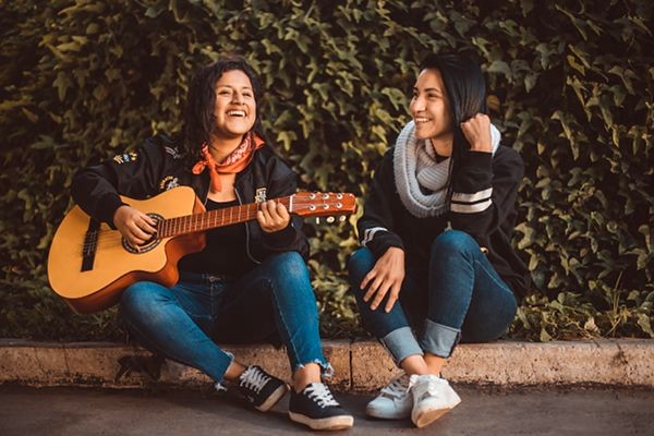 2 junge Frauen sitzen auf Randstein lachen Mädchen spielt Gitarre